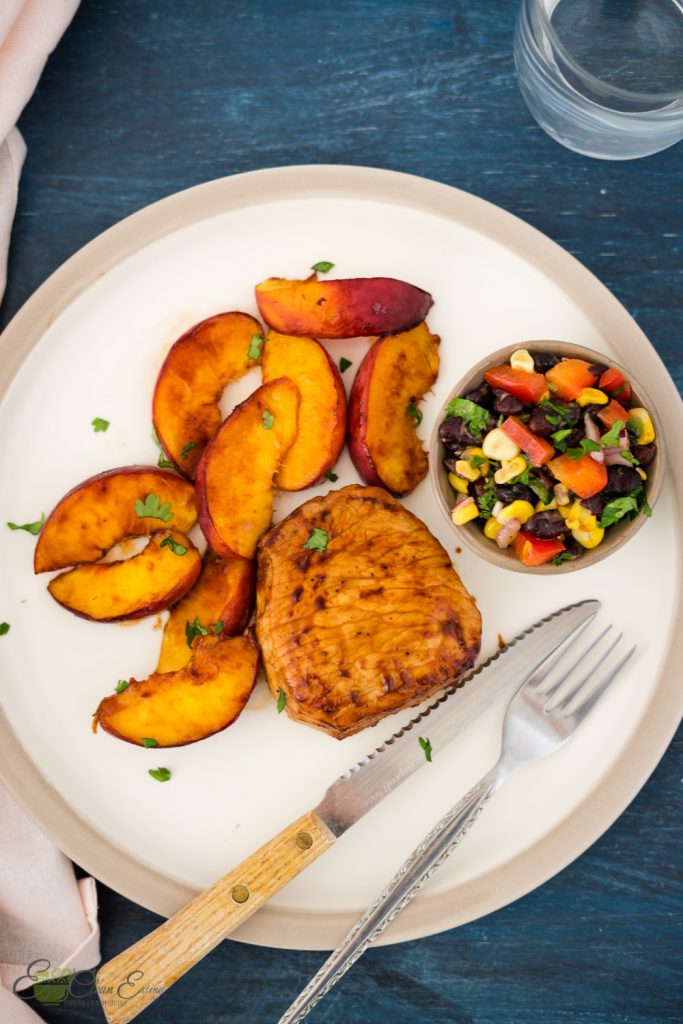 air fryer peaches with pork chops corn and black beans salsa, a fork and knife, water and napkin.