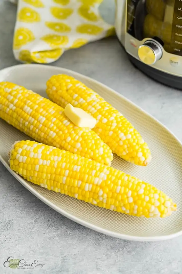 Perfectly cooked instant pot corn on the cob. with butter in a oval plate and  the pressure cooker behind.
