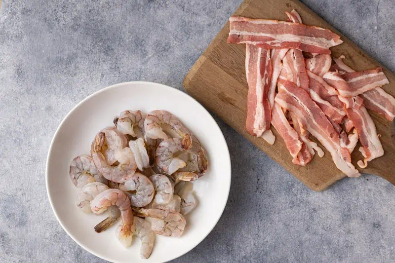 Raw shrimp in a bowl and bacon on a cutting board.