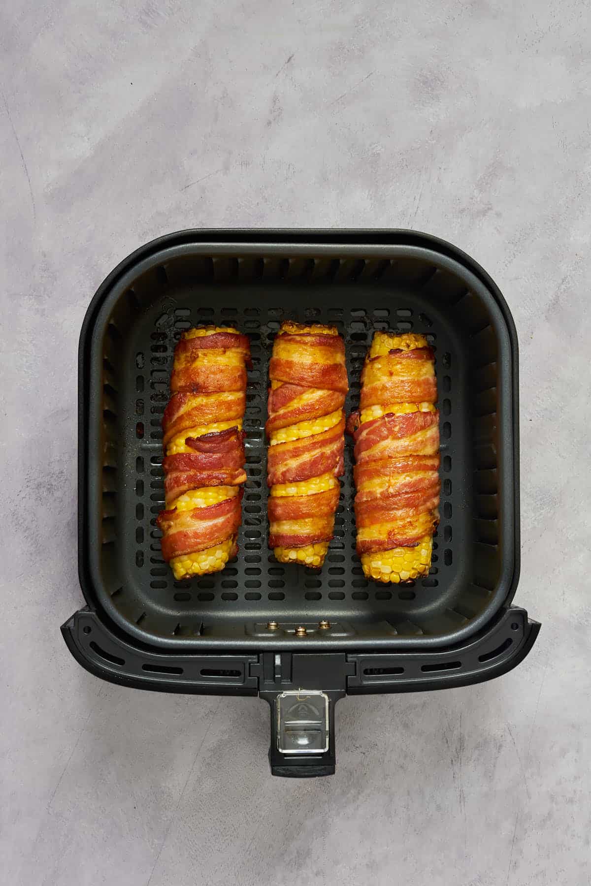 Inside the air fryer basket, corn on the cob wrapped in bacon after air frying.
