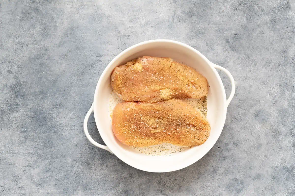 Uncooked chicken breasts with garlic powder, salt, and pepper in a bowl.