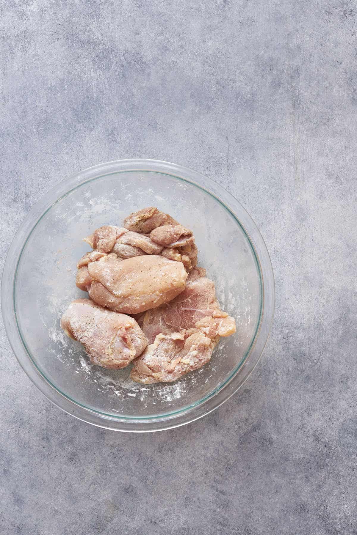 Chicken thighs seasoned with salt and pepper and coated with corn starch in a bowl.