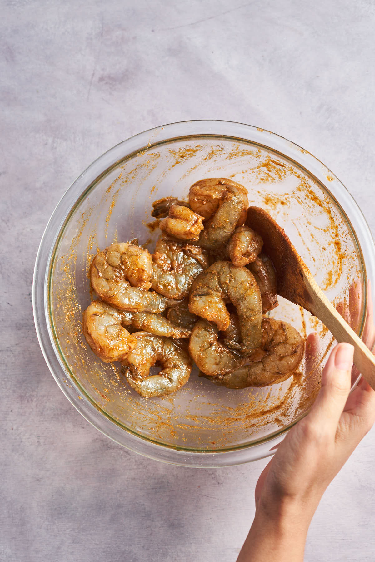 Shrimp being tossed with Cajun seasoning in a glass bowl using a wooden spoon.