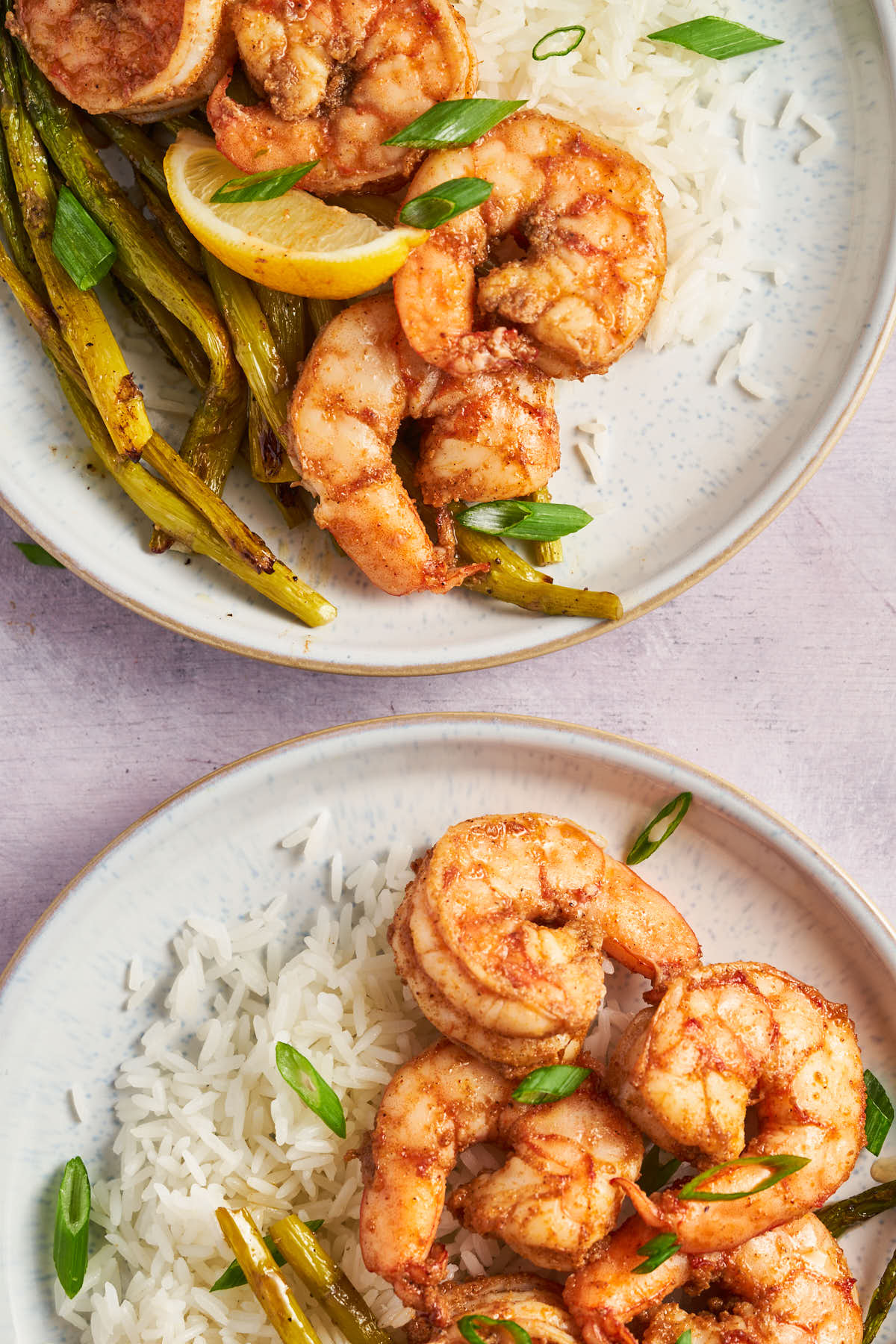 Two plates of Cajun shrimp with rice, roasted asparagus, and lemon wedges, viewed from above.