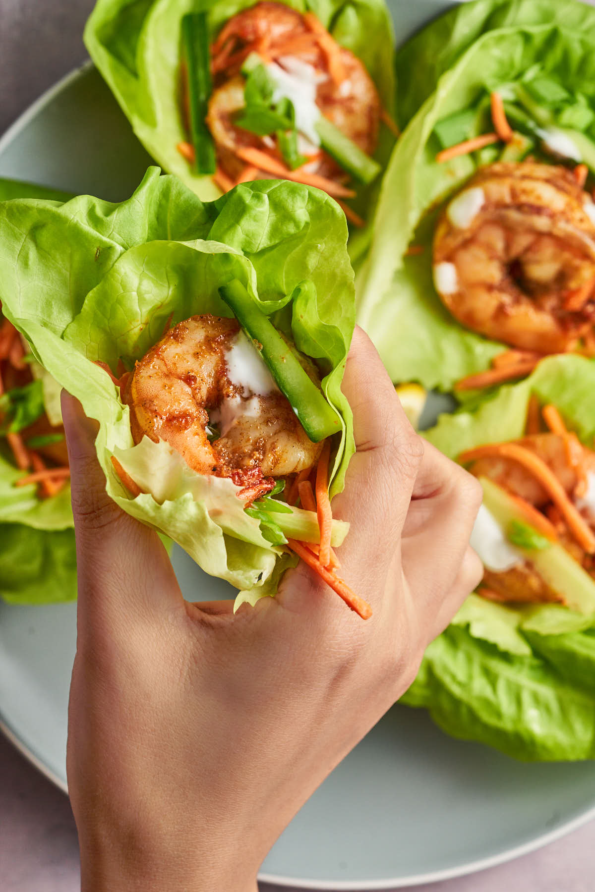 A hand holding a lettuce wrap filled with Cajun shrimp, cucumber, shredded carrots, and a creamy drizzle.