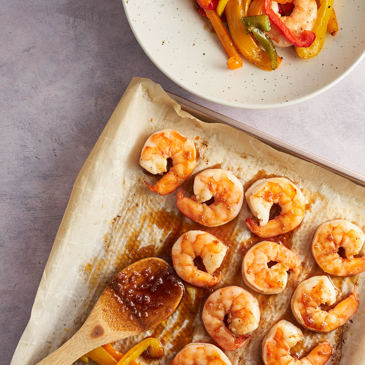 Honey garlic sauce on a wooden spoon beside cooked shrimp on a sheet pan.
