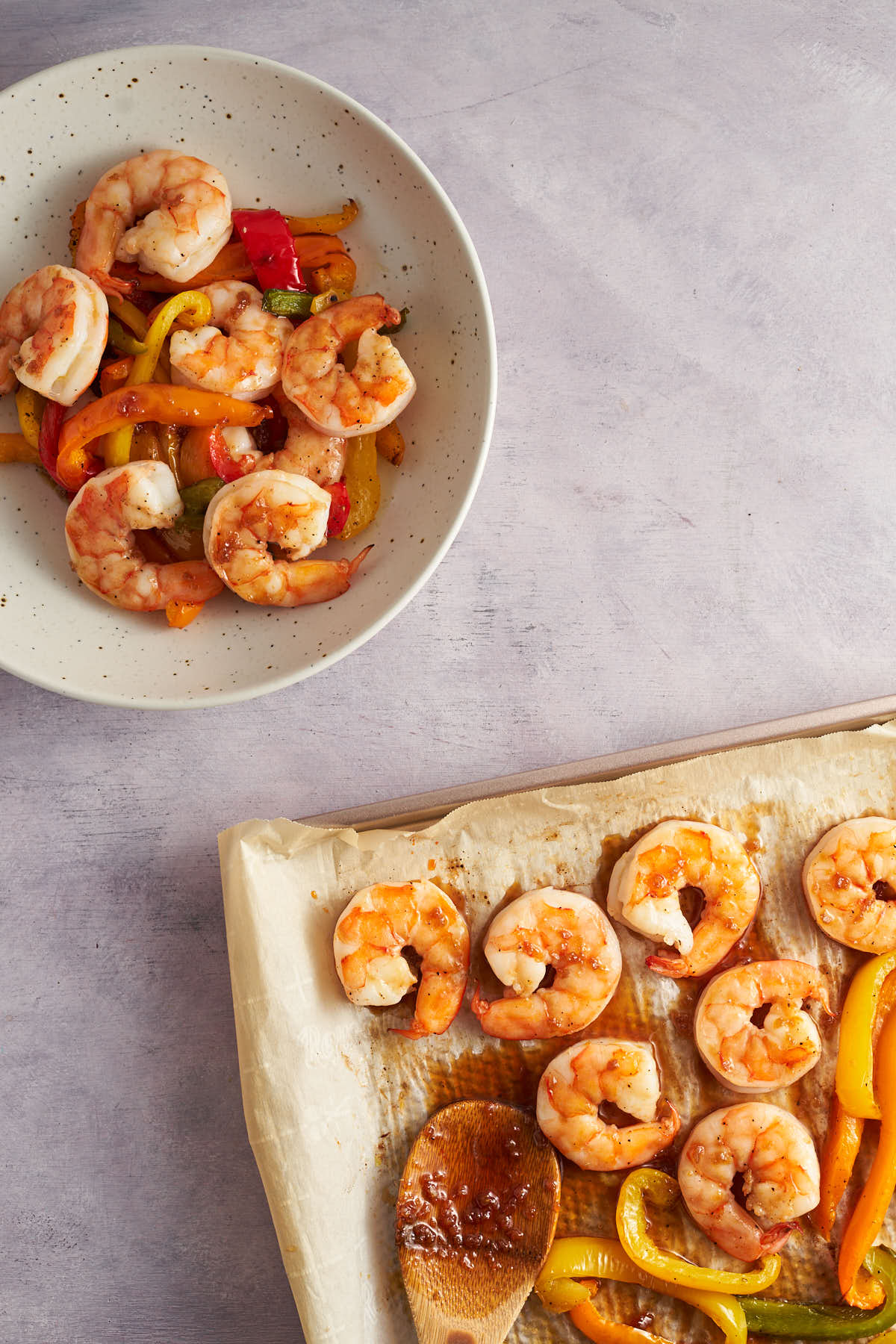 Honey garlic sauce on a wooden spoon beside cooked shrimp on a parchment lined sheet pan.