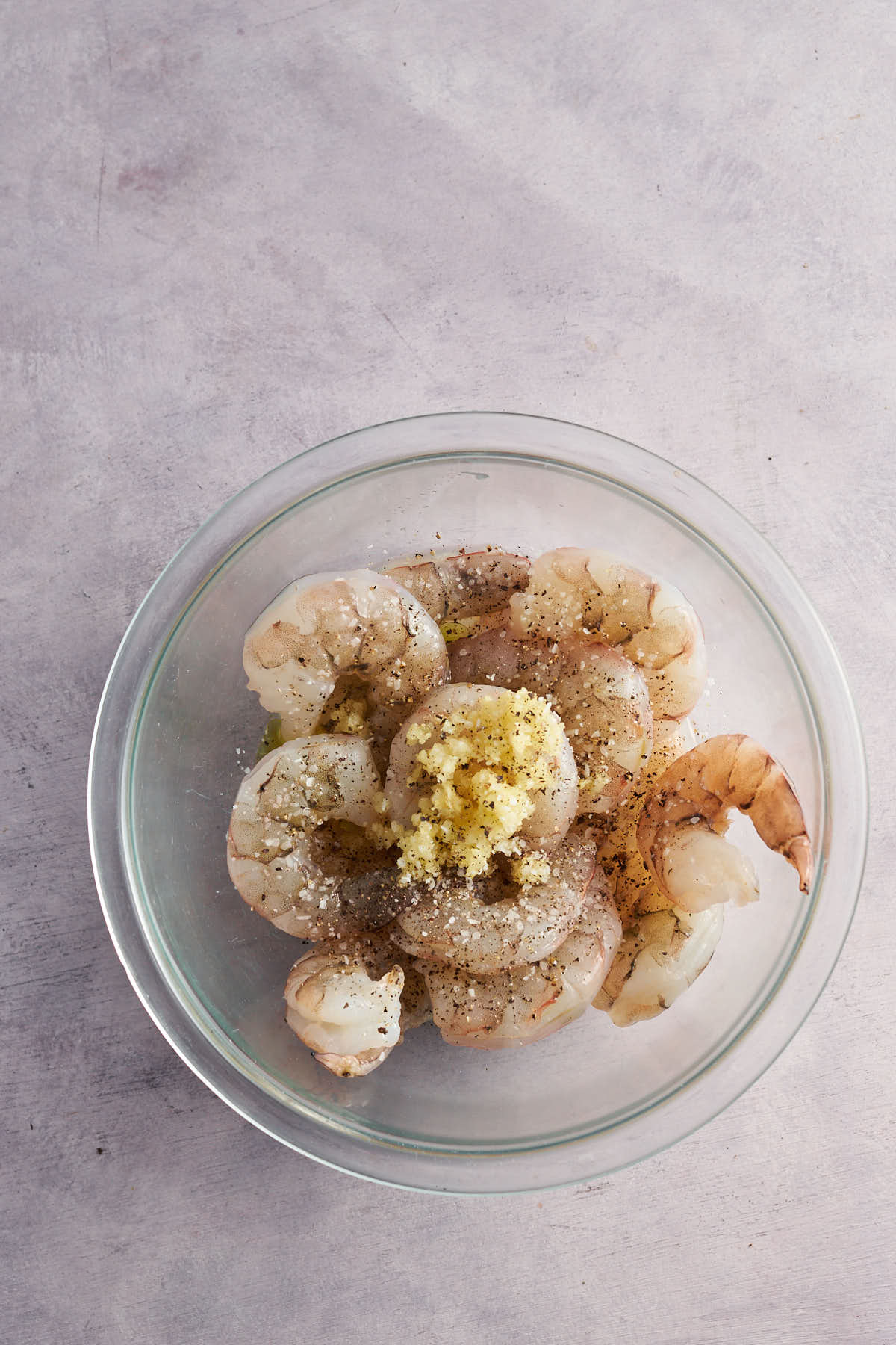 Raw shrimp topped with minced garlic and seasonings in a glass bowl.