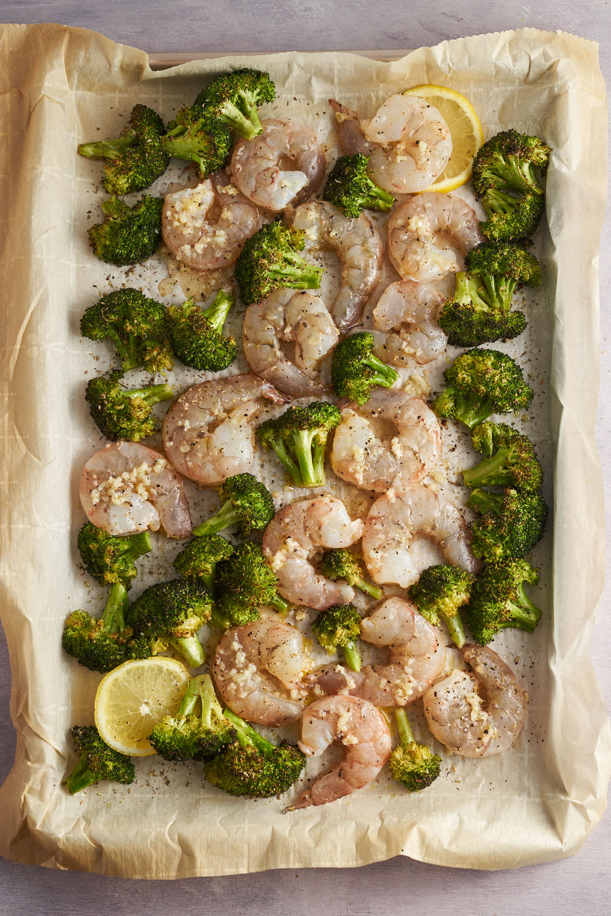 Raw shrimp and lemon slices arranged among roasted broccoli on a sheet pan.